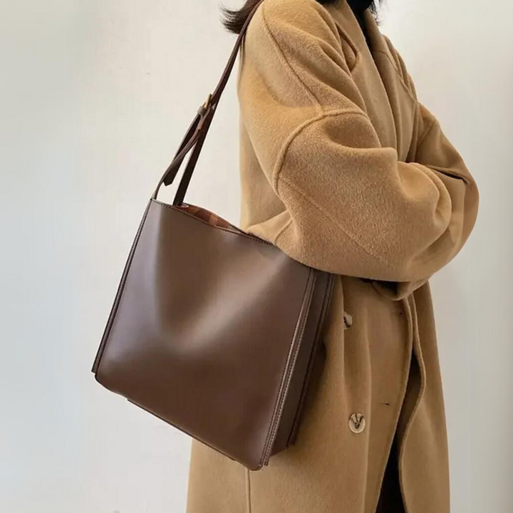 Damenhandtasche aus braunem Leder, getragen von einer Person im beigen Mantel. Stilvolle Umhängetasche, modisches Accessoire für den Alltag.