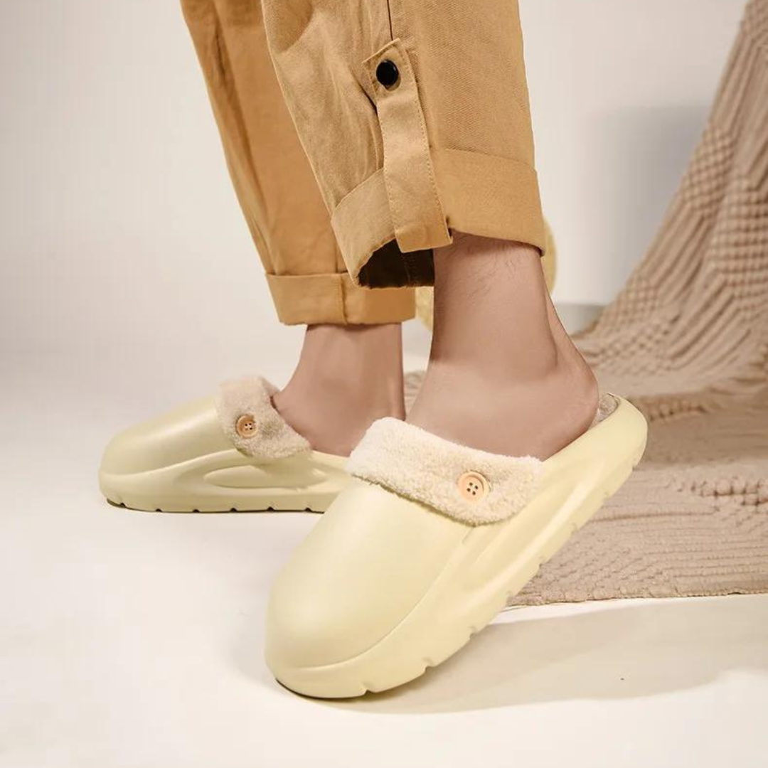 Beige Hausschuhe mit Fellfutter und Knopfdetail, getragen von Person in beigefarbener Hose. Gemütliche, warme Pantoffeln für den Winter.