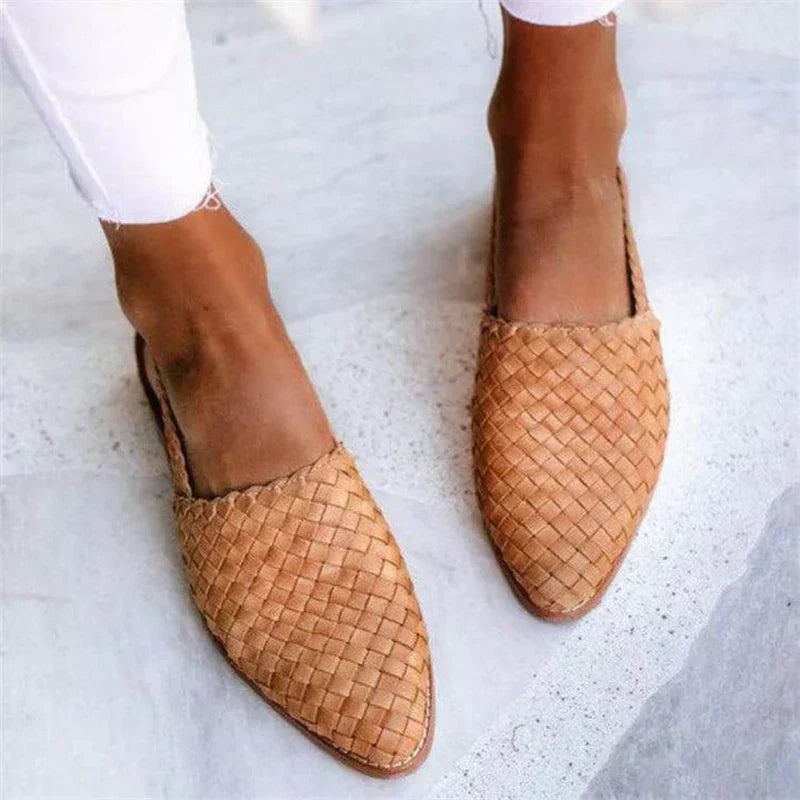 Braune geflochtene Lederslipper auf grauem Hintergrund, getragen von einer Person in weißen Hosen. Bequeme Sommerschuhe, handgefertigt, modisch.