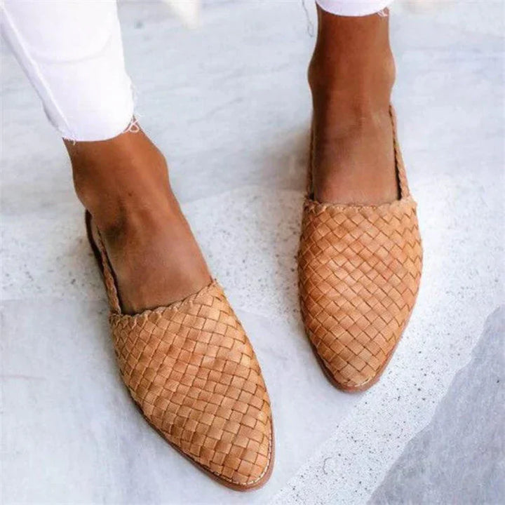 Braune geflochtene Lederslipper auf grauem Hintergrund, getragen von einer Person in weißen Hosen. Bequeme Sommerschuhe, handgefertigt, modisch.