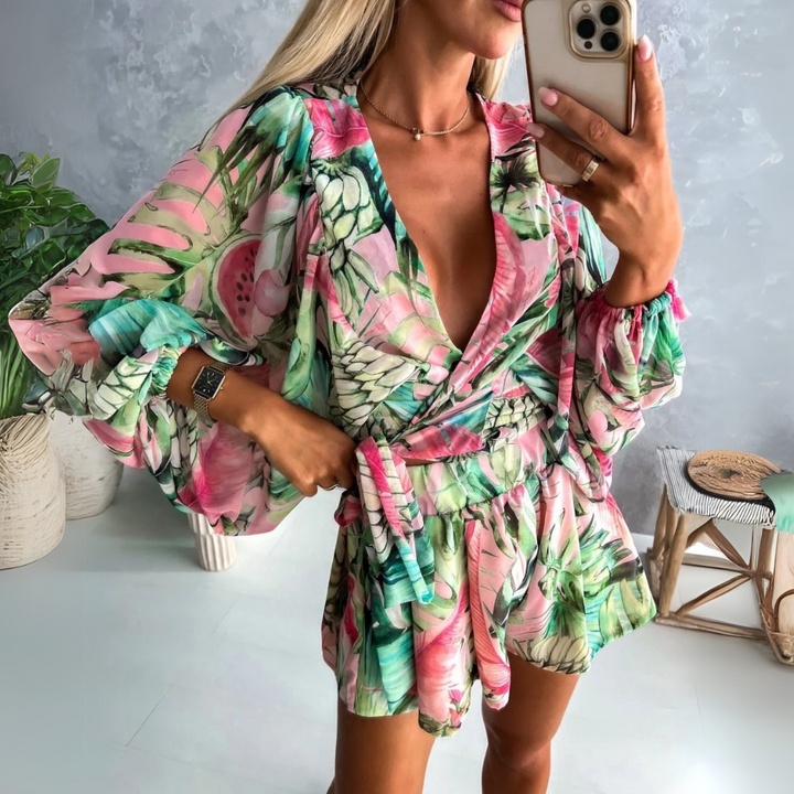 Frau in buntem, tropischem Jumpsuit mit Smartphone, modisches Sommeroutfit, helle Farben, Pflanzenmuster, moderne Inneneinrichtung im Hintergrund.