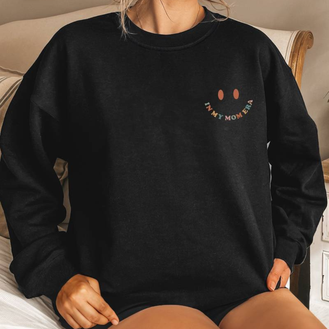 Frau trägt schwarzen Pullover mit Smiley und "In My Mom Era"-Aufdruck, sitzt auf Bett. Bequemes, lässiges Outfit, ideal für Freizeitmode.