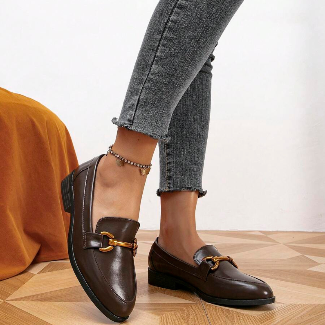 Damen braune Loafer mit goldener Schnalle, kombiniert mit grauen Jeans. Modische Schuhe auf Holzboden, ideal für eleganten Casual-Look.