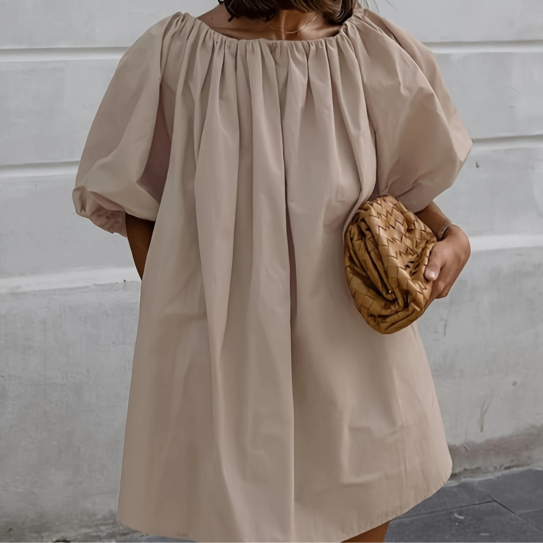 Frau in beigem, schulterfreiem Kleid mit Puffärmeln hält geflochtene Clutch. Elegantes Sommeroutfit, modisch und stilvoll.