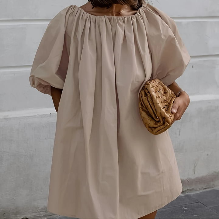 Frau in beigem, schulterfreiem Kleid mit Puffärmeln hält geflochtene Clutch. Elegantes Sommeroutfit, modisch und stilvoll.