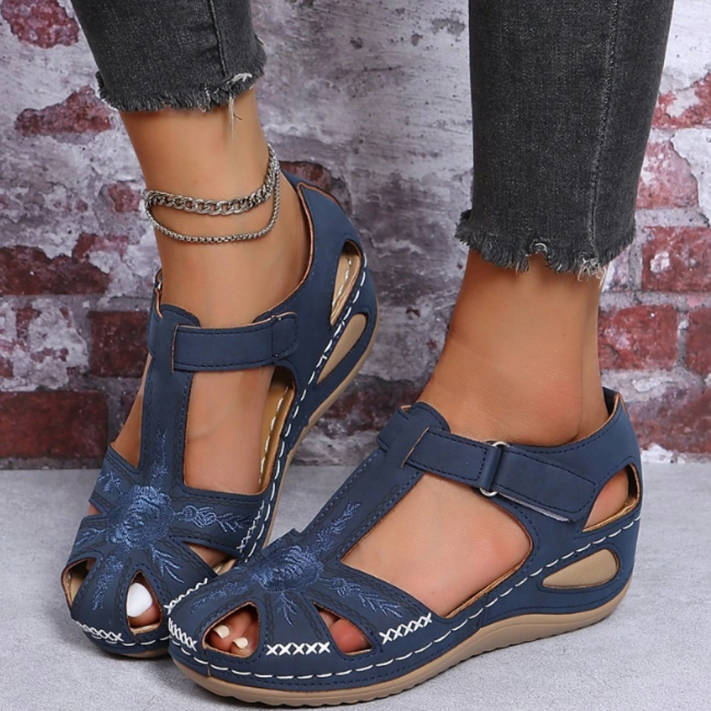 Damen Sandalen in Blau mit Klettverschluss, gesticktem Blumenmuster und Keilabsatz, getragen mit grauen Jeans vor Backsteinwand.