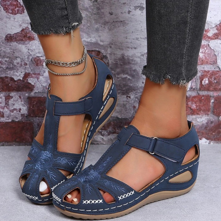 Damen Sandalen in Blau mit Klettverschluss, gesticktem Blumenmuster und Keilabsatz, getragen mit grauen Jeans vor Backsteinwand.