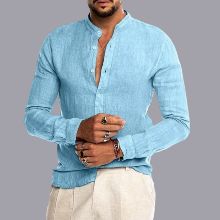 Mann in hellblauem Leinenhemd mit Stehkragen, trägt mehrere Ringe und Armbänder, vor grauem Hintergrund. Herrenmode, Freizeitlook, Sommeroutfit.