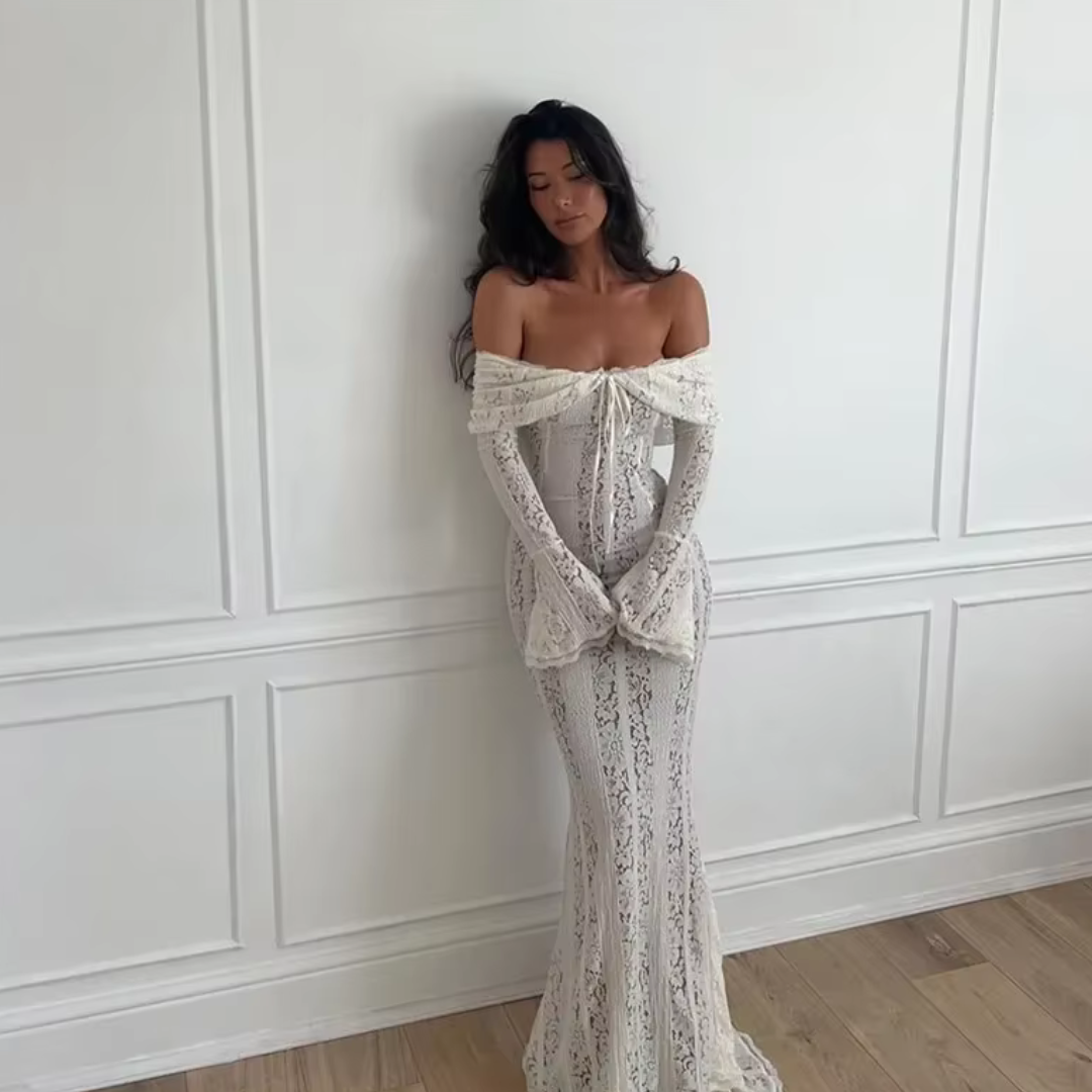 Frau in eleganter, schulterfreier Spitzenkleid, posiert vor weißer Wand. Hochzeitsmode, Brautkleid, stilvolle Abendgarderobe.