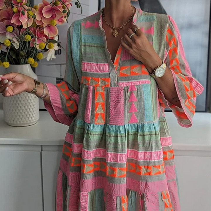 Frau in buntem, geometrisch gemustertem Kleid vor Blumenstrauß. Sommermode, Boho-Stil, leuchtende Farben, Freizeitkleidung, modisches Outfit.