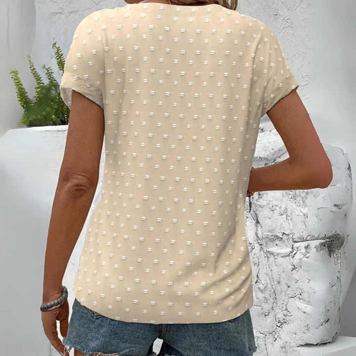 Rückansicht einer Frau in beigem, gepunktetem T-Shirt und Jeansshorts, vor weißer Wand mit Pflanzen. Sommermode, lässiger Stil, Damenbekleidung.