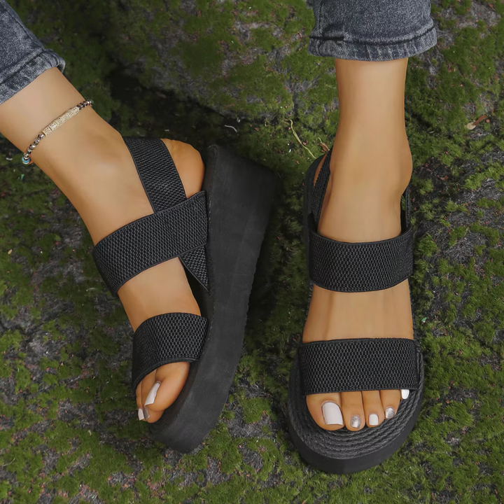 Schwarze Plateau-Sandalen mit breiten Riemen an weiblichen Füßen, getragen auf moosbedecktem Boden. Modische Sommerschuhe, bequem und stilvoll.