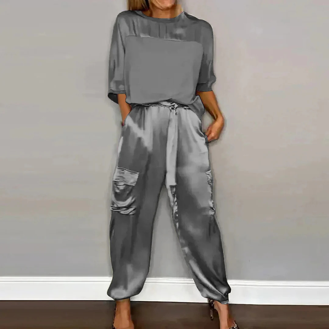 Frau in grauem Satin-Zweiteiler mit lockerem Oberteil und Jogginghose, modisch, elegant, lässig, vor grauer Wand, moderne Damenmode.