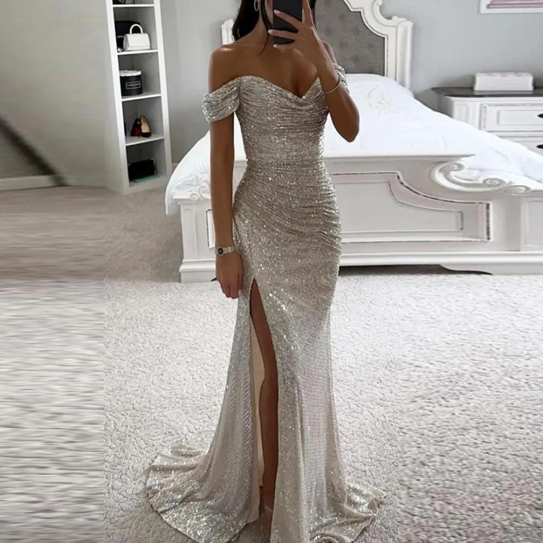 Elegantes, schulterfreies Abendkleid in Silber mit Pailletten und hohem Beinschlitz, getragen von einer Frau in einem stilvollen Schlafzimmer.