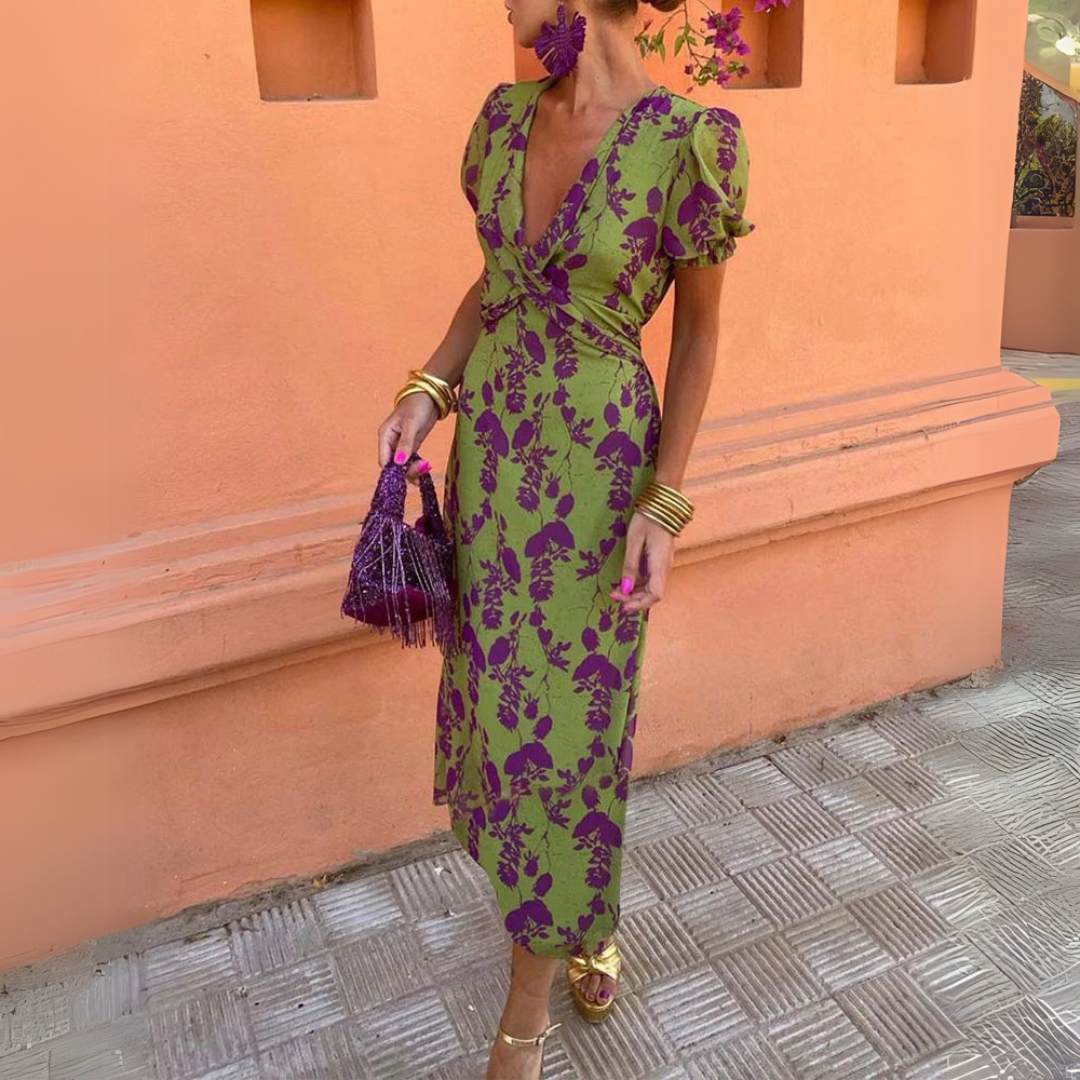 Frau in grünem, geblümtem Kleid mit lila Akzenten, hält eine lila Tasche, steht vor orangefarbener Wand. Elegante Mode, Sommerkleid, Streetstyle.