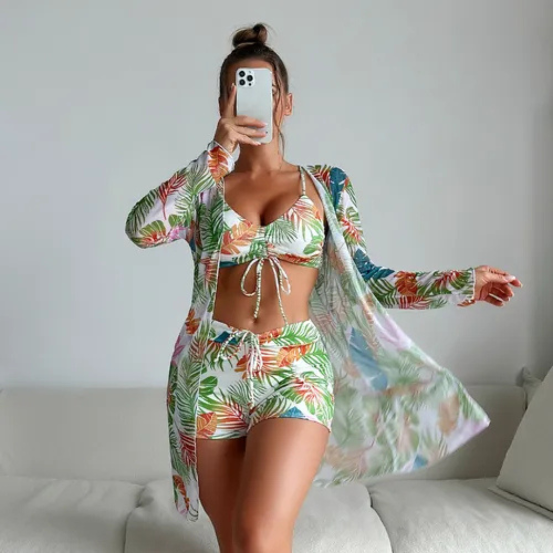 Frau in tropischem Bikini-Set mit passendem Kimono, macht Selfie. Sommermode, Strandoutfit, tropisches Muster, Handy, Innenraum.