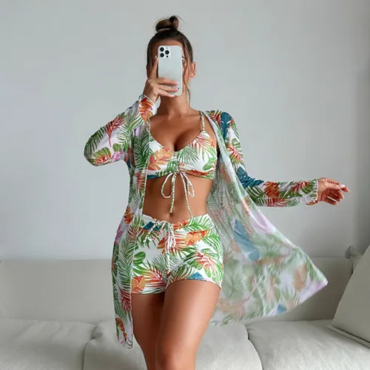 Frau in tropischem Bikini-Set mit passendem Kimono, macht Selfie. Sommermode, Strandoutfit, tropisches Muster, Handy, Innenraum.