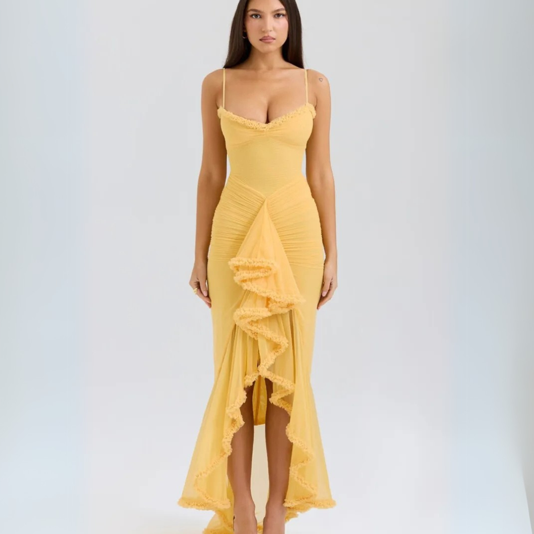 Frau in gelbem, figurbetontem Abendkleid mit Rüschen, Spaghettiträgern und asymmetrischem Saum, vor neutralem Hintergrund. Elegante Mode.