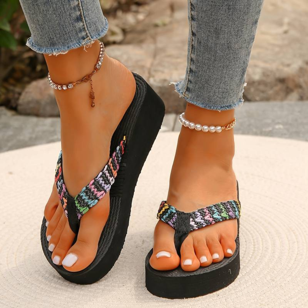 Frau trägt bunte Plateau-Flip-Flops und Jeans, Schmuck an den Knöcheln, auf Steinboden. Sommermode, Fußbekleidung, Freizeitlook.