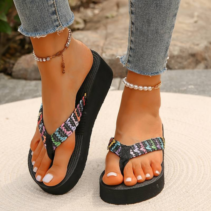 Frau trägt bunte Plateau-Flip-Flops und Jeans, Schmuck an den Knöcheln, auf Steinboden. Sommermode, Fußbekleidung, Freizeitlook.