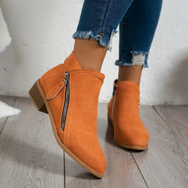 Orange Damen-Stiefeletten mit Reißverschluss, kombiniert mit zerrissenen Jeans auf Holzboden. Modische Herbstschuhe, bequeme Absatzstiefeletten.