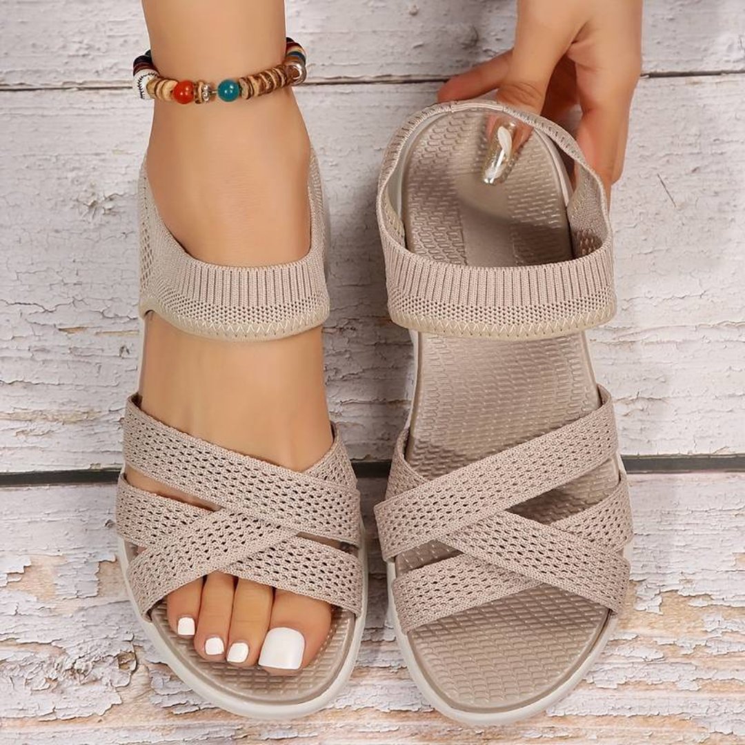 Beige Damen-Sandalen mit Kreuzriemen, gestricktem Design und Fußkettchen auf Holzuntergrund. Sommermode, bequeme Schuhe, trendige Accessoires.