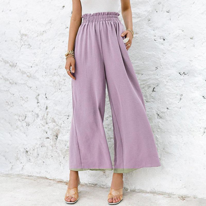 Frau in lila weiten Hosen vor weißer Wand, modische Sommerkleidung, lässiger Stil, elegante Sandalen, trendige Damenmode.