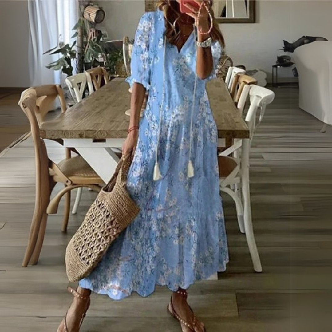 Frau in blauem Blumenkleid mit Strohtasche steht in modernem Esszimmer mit Holztisch und Stühlen. Sommermode, Inneneinrichtung, Boho-Stil.