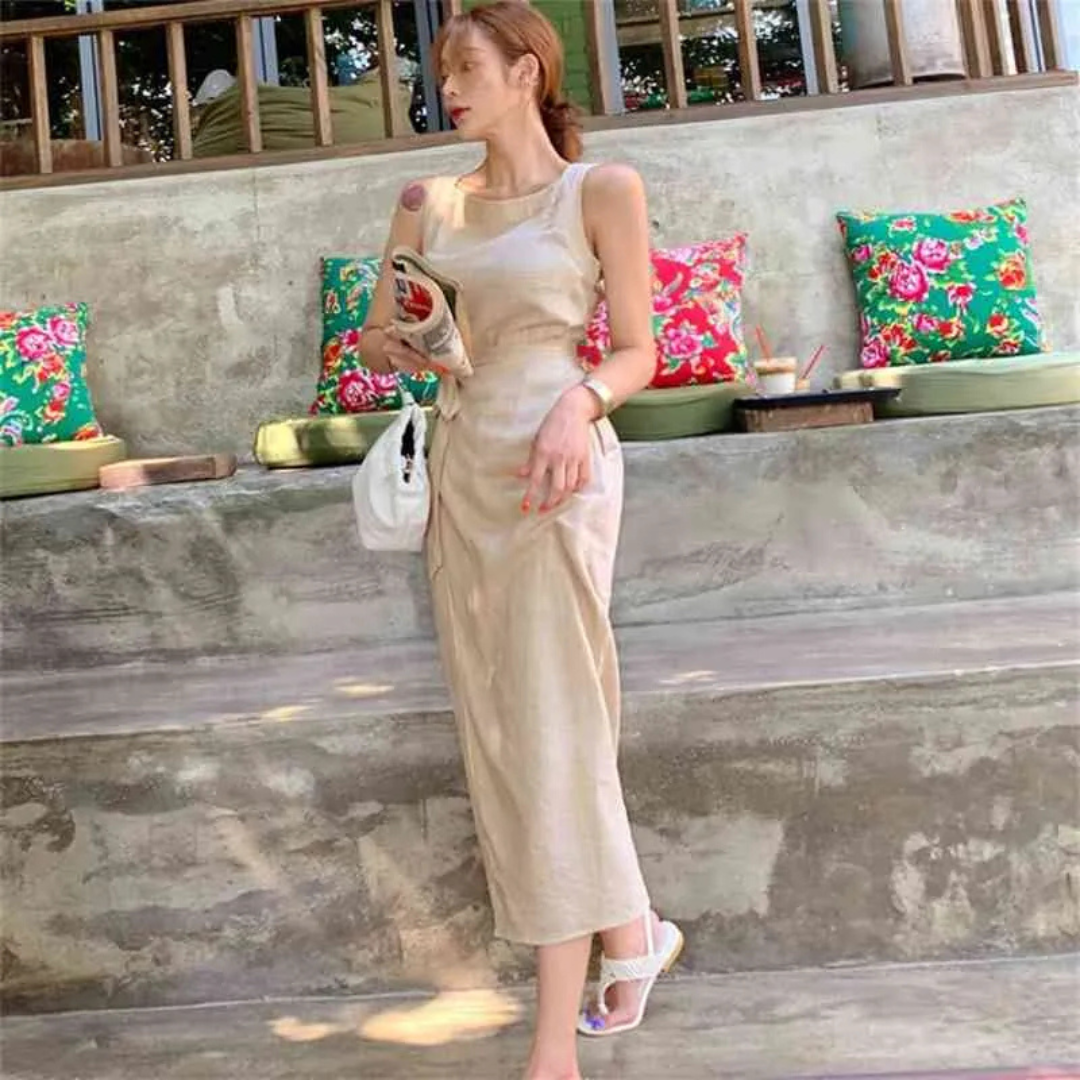 Frau in beigem Sommerkleid mit Handy und weißer Tasche, steht vor einer Steinmauer mit bunten Kissen. Perfektes Outfit für Sommermode und Freizeit.