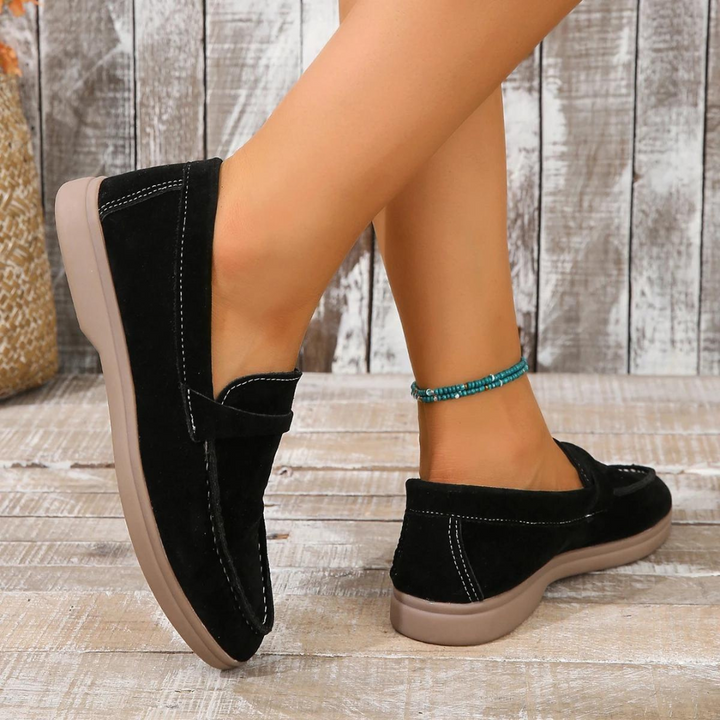 Schwarze Damen-Loafer aus Wildleder mit bequemer Sohle, getragen von einer Person mit türkisfarbenem Fußkettchen, auf Holzboden.
