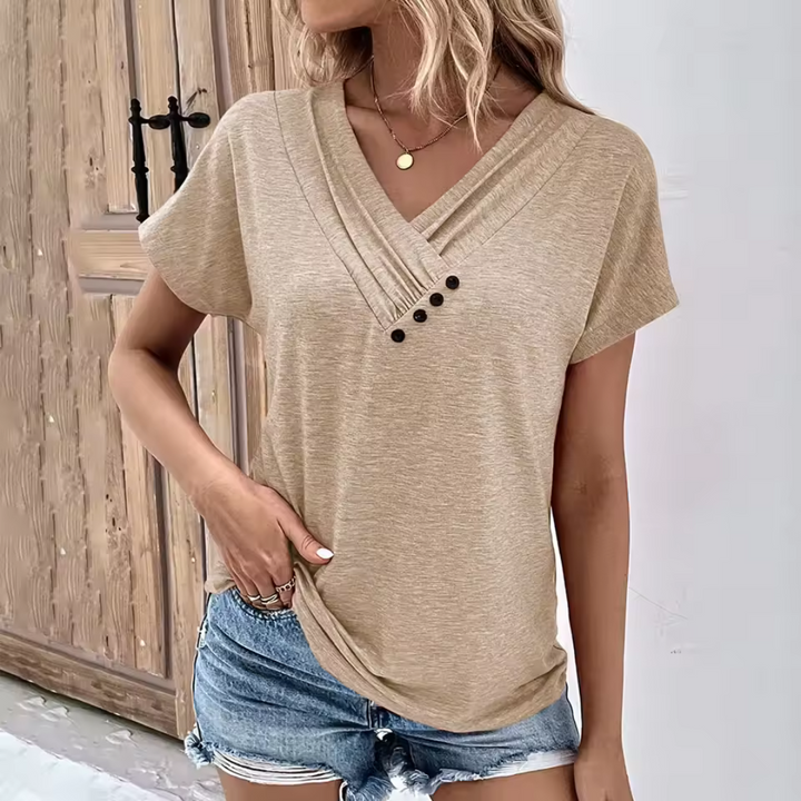 Blonde Frau trägt beige T-Shirt mit Knopfleiste und blaue Jeansshorts, steht vor Holztür. Sommermode, lässiger Stil, Damenbekleidung.