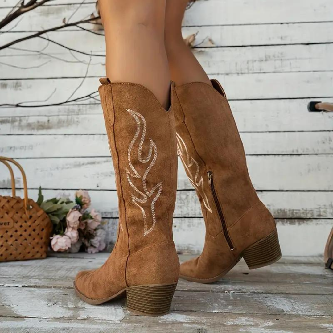 Braune Wildleder-Cowboystiefel mit Stickerei, niedriger Absatz, seitlicher Reißverschluss, vor weißer Holzwand, modisch und trendig.