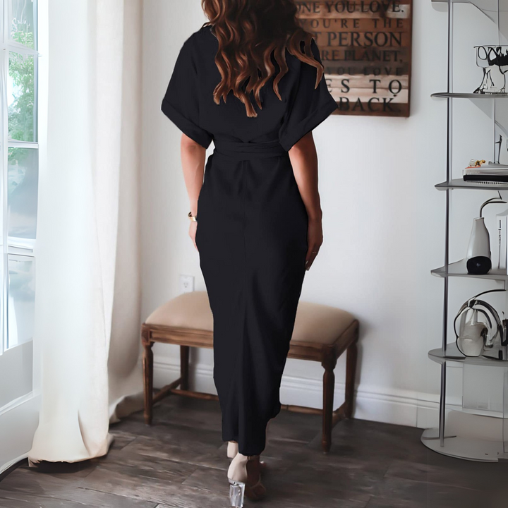 Frau in elegantem schwarzen Kleid, Rückansicht, steht in modernem Wohnzimmer mit Bücherregal und Bank, stilvolle Inneneinrichtung, Tageslicht.