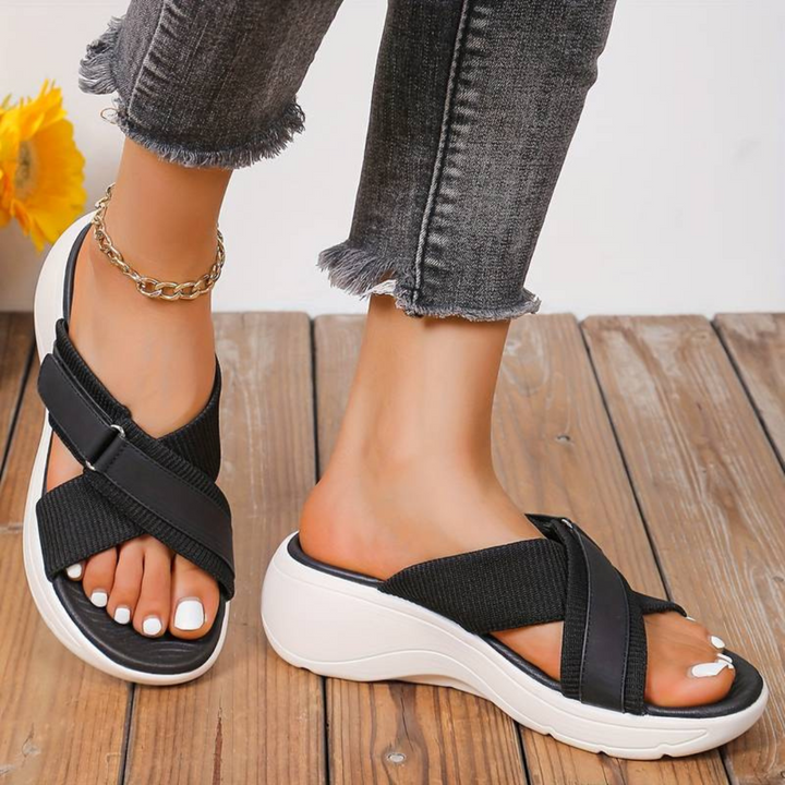 Schwarze Damen-Sandalen mit Keilabsatz und Kreuzriemen, getragen auf Holzterrasse. Modische Sommerschuhe, bequem und stilvoll.