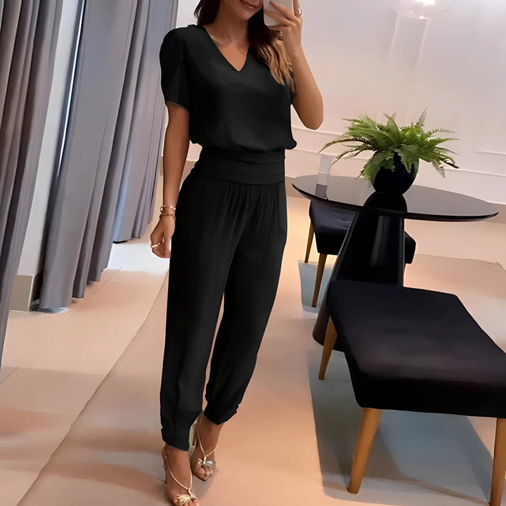 Frau in eleganter schwarzer Kleidung posiert in modernem Wohnzimmer mit Tisch und Pflanzen, trägt goldene Sandalen. Stilvolle Mode, Inneneinrichtung.