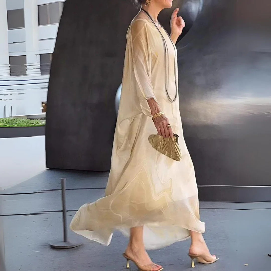 Elegante Frau in fließendem, cremefarbenem Maxikleid und High Heels, trägt goldene Clutch und Schmuck, modernes urbanes Umfeld im Hintergrund.