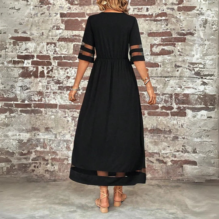 Frau in langem, schwarzem Kleid mit transparenten Ärmeln vor einer Backsteinwand. Elegantes Abendkleid, modisches Design, stilvolle Damenmode.