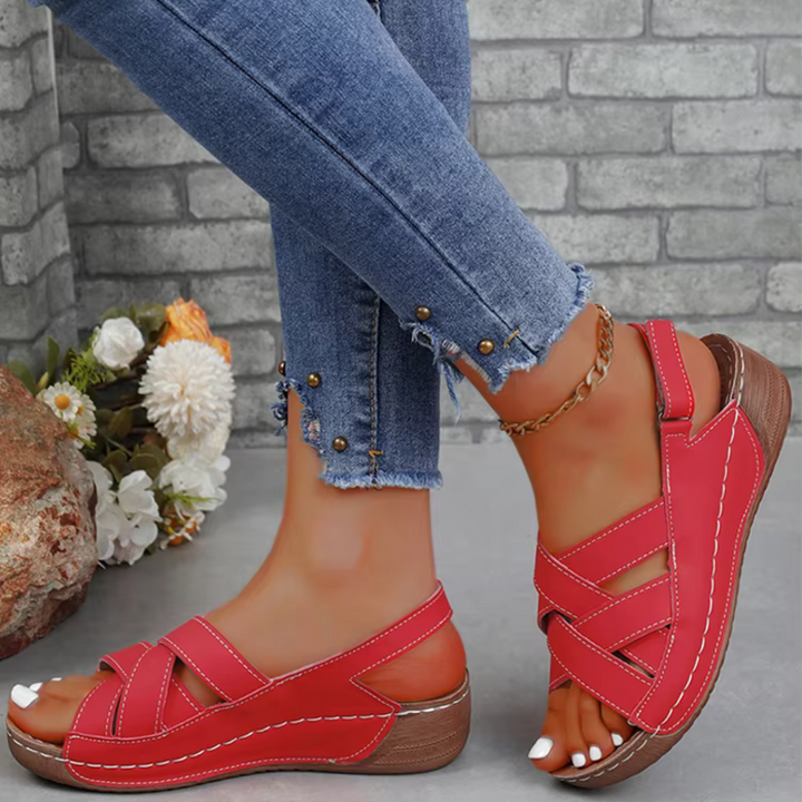Rote Sandalen mit Keilabsatz an Frauenfüßen, kombiniert mit blauen Jeans mit Perlendetails, vor einer Ziegelwand und Blumen im Hintergrund.