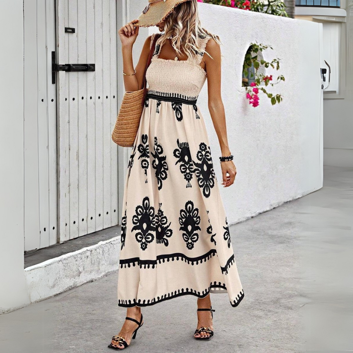 Frau in langem, ärmellosem Sommerkleid mit schwarzem Muster, Strohhut und Sandalen, steht vor weißer Wand. Sommermode, Boho-Stil, Strandkleidung.