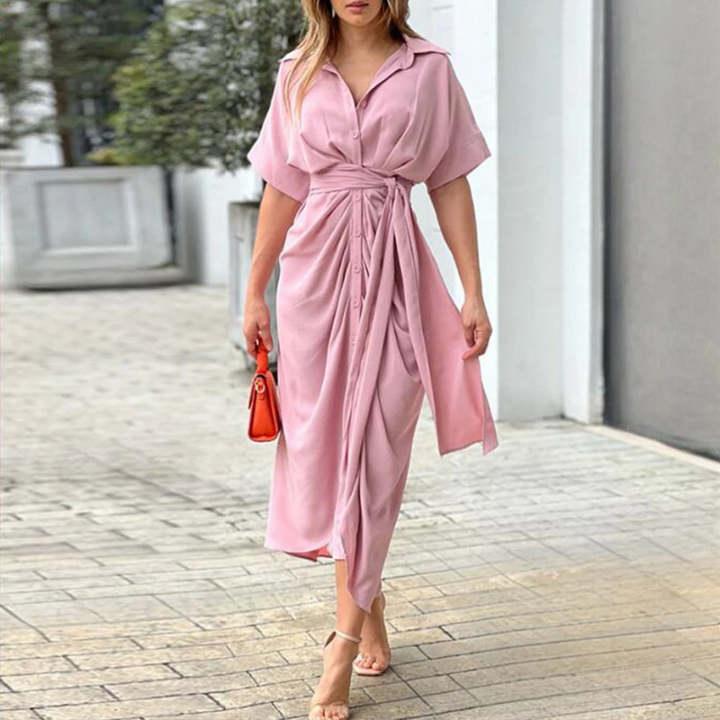 Frau in elegantem rosa Wickelkleid mit kurzen Ärmeln, trägt rote Handtasche und Sandalen, läuft auf gepflastertem Weg. Mode, Damenkleidung, Sommeroutfit.
