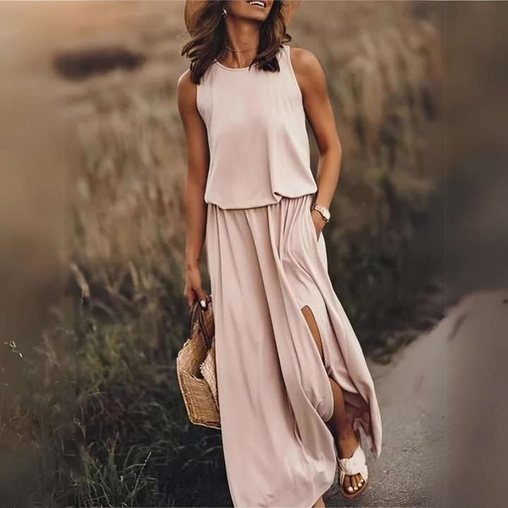 Frau in einem eleganten, ärmellosen Maxikleid in Beige, mit Strohhut und Korbtasche, spaziert auf einem ländlichen Weg. Sommermode, Freizeitlook.