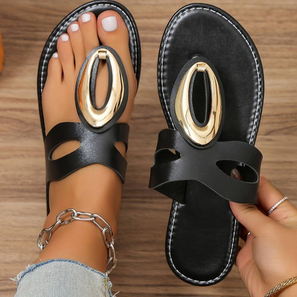 Schwarze Sandalen mit goldener Verzierung auf Holzuntergrund, modisch, Damenfuß mit Fußkettchen, Sommermode, stilvolle Schuhe.