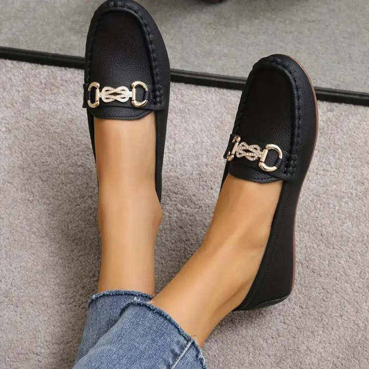 Schwarze Damen-Loafer mit goldener Schnalle, getragen auf grauem Teppich. Modische, bequeme Schuhe, ideal für Freizeit und Büro.