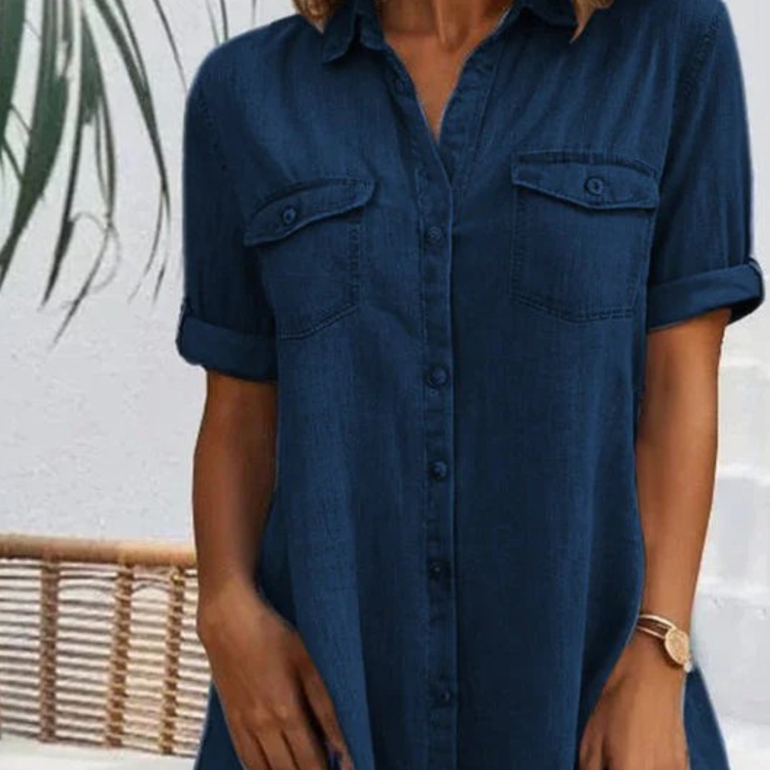 Frau in dunkelblauem Jeanskleid mit aufgerollten Ärmeln, vor weißer Wand und Pflanzen. Modetrend, lässiger Sommerlook, Damenbekleidung.