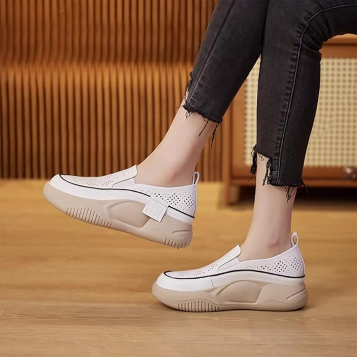 Weiße Damen-Sneaker mit dicker Sohle, perforiertem Design und Klettverschluss, getragen zu schwarzen Jeans. Bequeme, modische Freizeitschuhe.