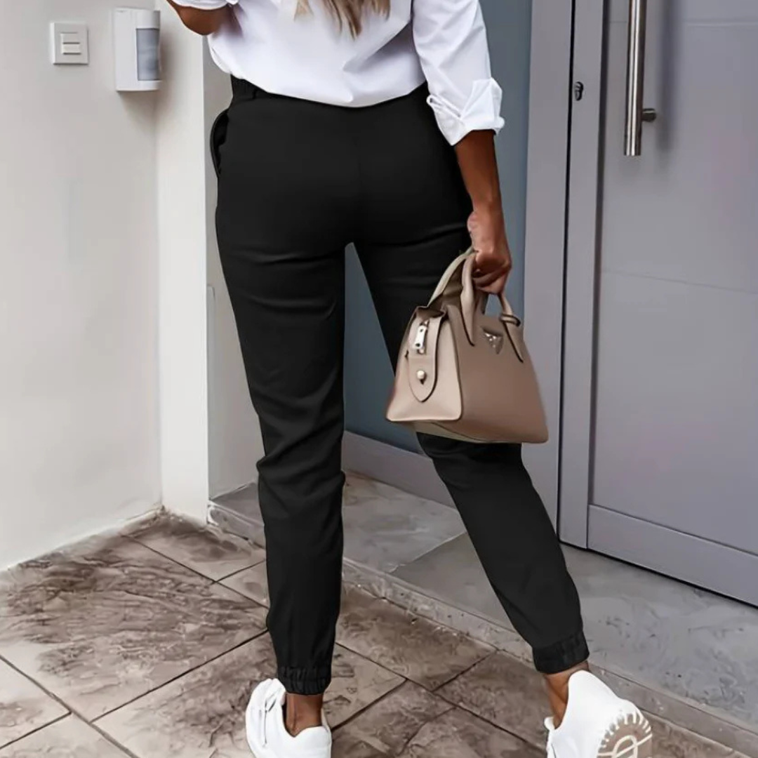 Frau in schwarzer Hose und weißem Hemd, hält beige Handtasche, trägt weiße Sneaker, steht vor moderner Haustür. Mode, Streetstyle, Damenbekleidung.