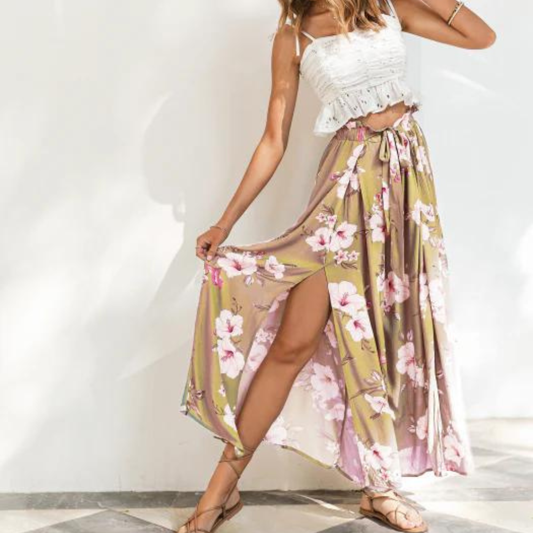 Frau in weißem Spitzenoberteil und geblümtem Maxirock posiert vor weißer Wand. Sommermode, Boho-Stil, florales Design, luftiges Outfit.