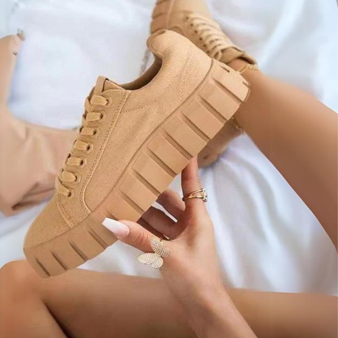 Beige Plateau-Sneaker mit dicker Sohle, gehalten von einer Hand mit weiß lackierten Nägeln, auf weißem Hintergrund. Trendige Damenmode, Schuhe.