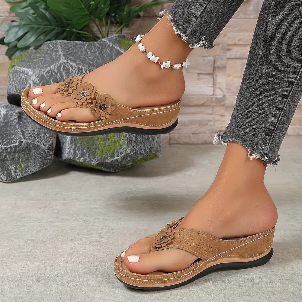 Damenfüße in braunen Keilsandalen mit Blumenmuster, Jeans mit Fransen, auf grauem Boden vor Steinwand und Pflanzen. Modische Sommerschuhe.