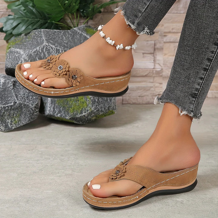 Damenfüße in braunen Keilsandalen mit Blumenmuster, Jeans mit Fransen, auf grauem Boden vor Steinwand und Pflanzen. Modische Sommerschuhe.
